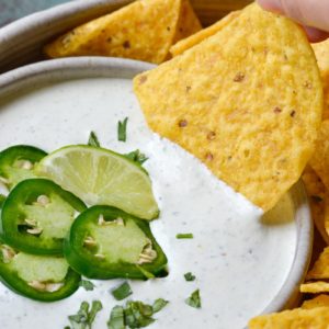 tortilla chip being dipped into a bowl of creamy ranch dip topped with jalapenos