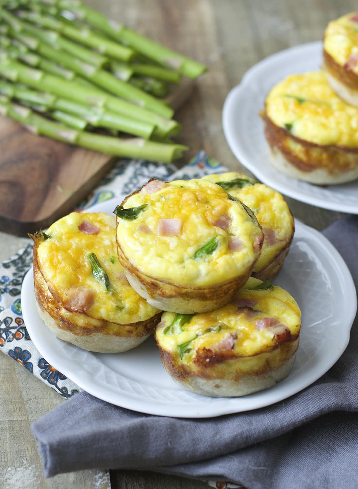 A stack of four ham and asparagus quiche on a white plate. Fresh asparagus spears rest in the background, along with another plate of mini quiche.
