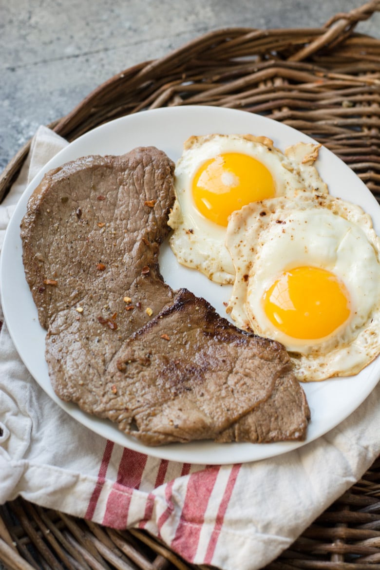 breakfast steak and eggs on a white plate. The plate is on a tea towel, which is on a wicker tray 