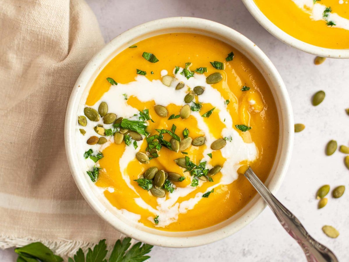 horizontal image showing a white bowl of butternut squash soup with a swirl of cream and sprinkled with fresh herbs and pumpkin seeds