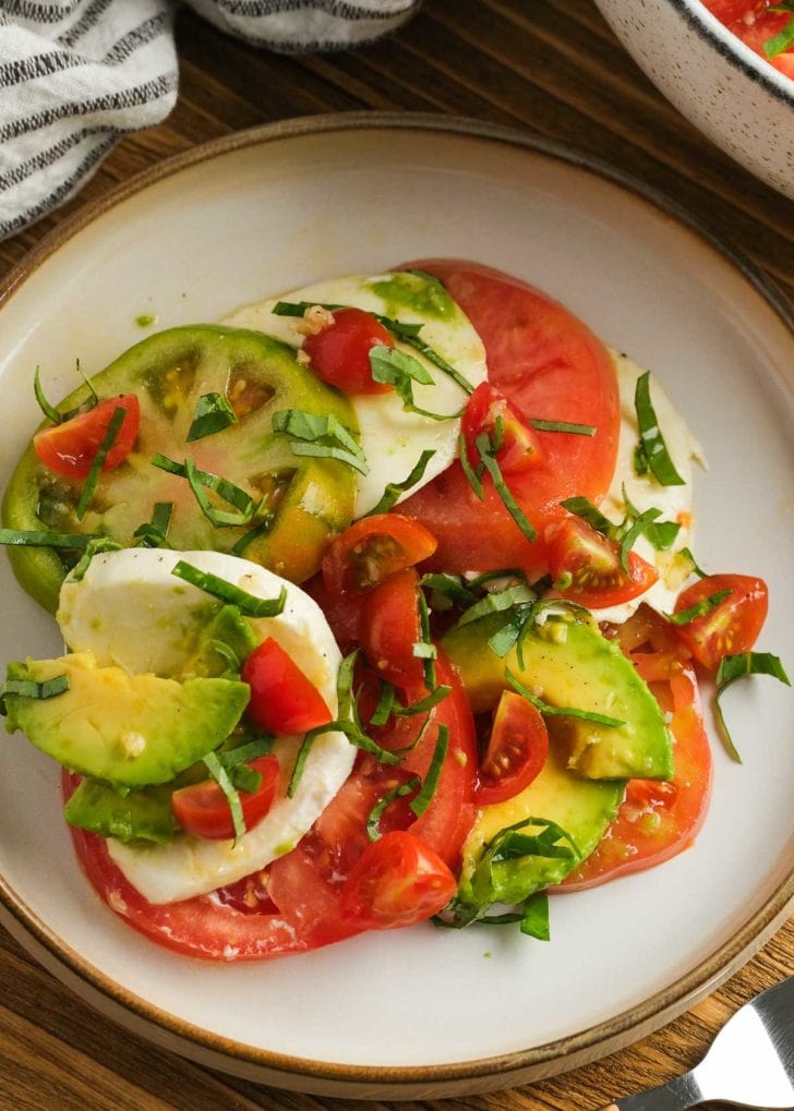 a plate with a well-dressed tomato salad with avocado, mozzarella, and basil