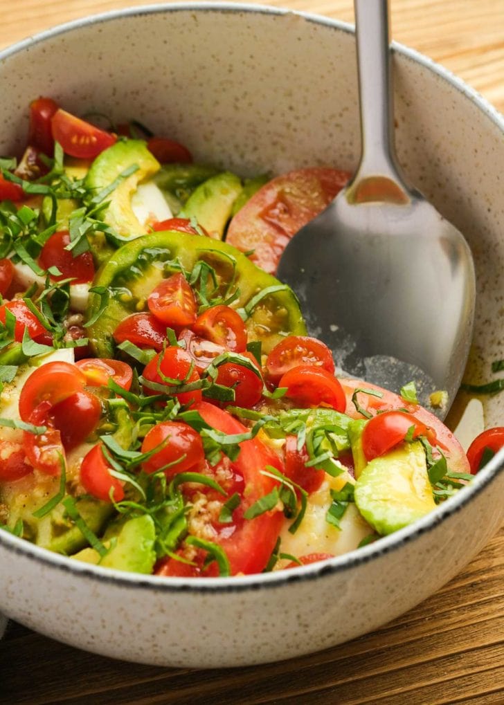 a large spoon in a white bowl full of tomato and avocado salad with mozzarella and basil