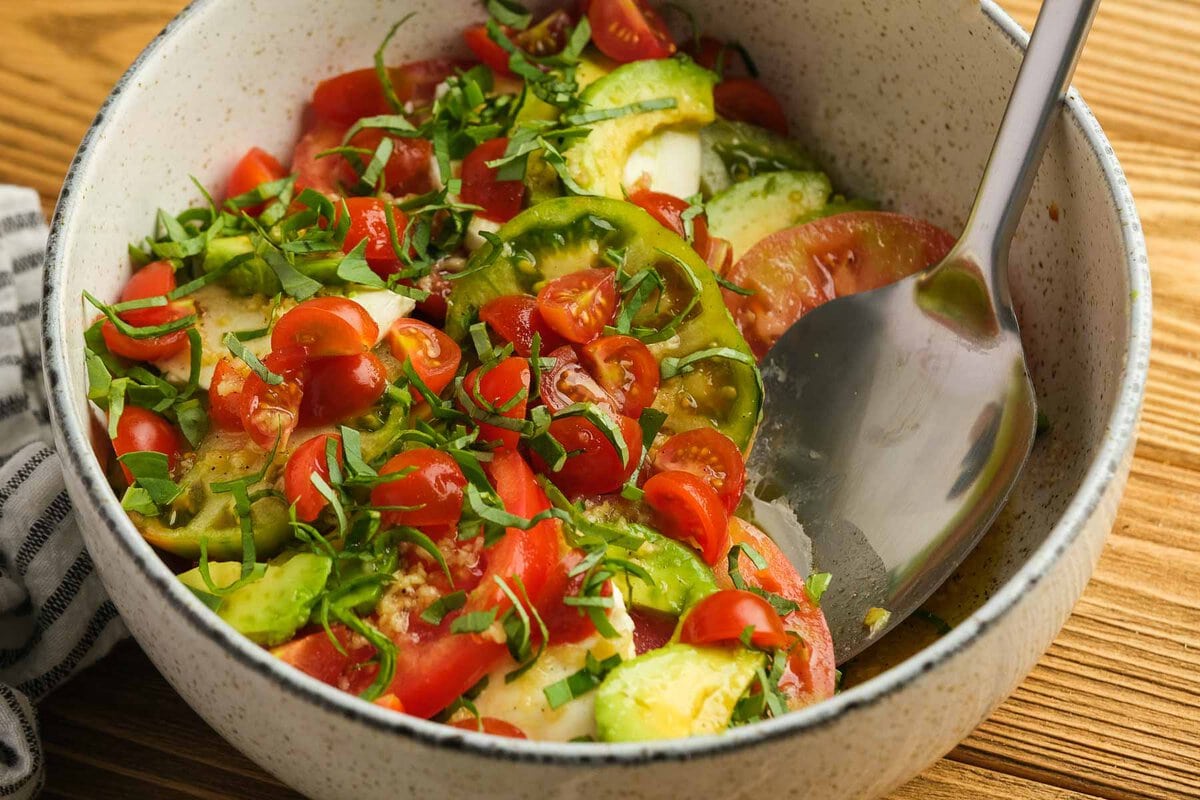 closeup view of a spoon scooping a serving of salad with tomatoes, avocados, basil, and mozzarella