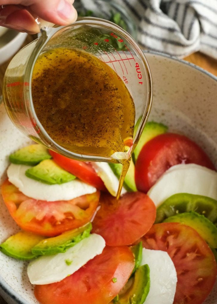 homemade vinaigrette being drizzled over tomato, avocado, and mozzarella slices in a bowl