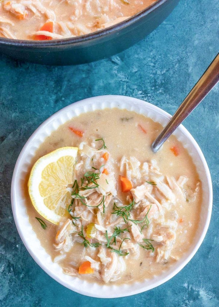 overhead shot showing a white bowl of rich avgolemono soup topped with fresh herbs and a lemon wedge