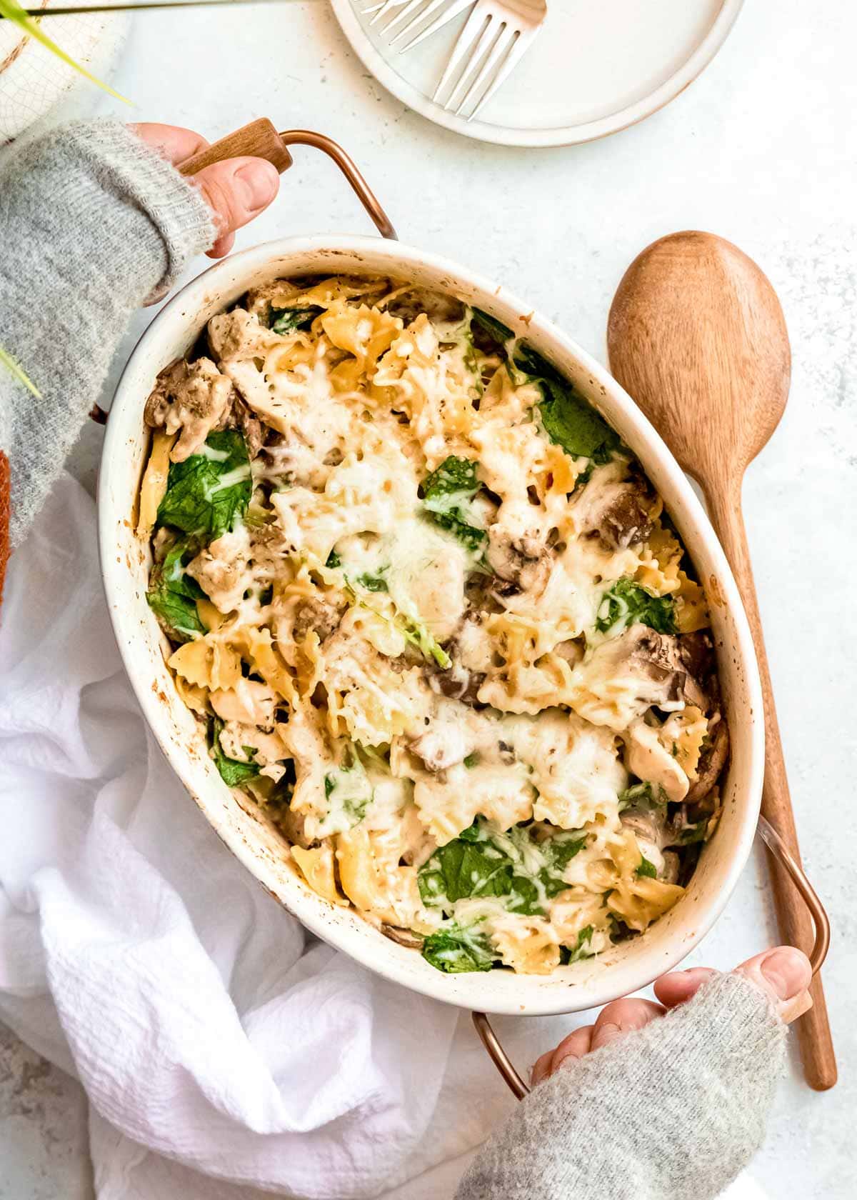 overhead shot showing hands placing a large casserole dish full of baked chicken and mushroom pasta topped with creamy cheese onto the table next to a wooden spoon