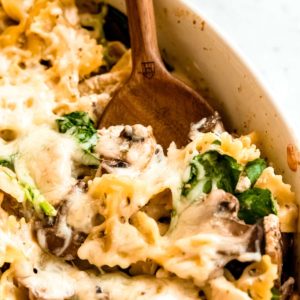 baked chicken and mushroom pasta being scooped out of white baking dish with wooden spoon