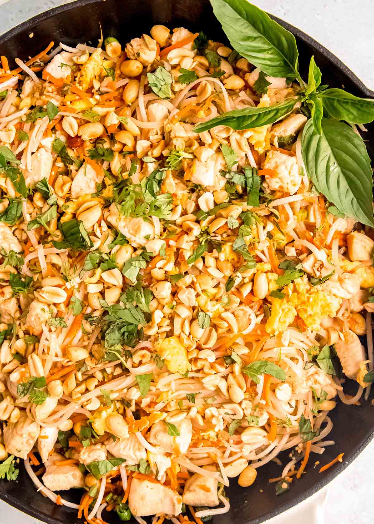 a wok full of tender pad thai noodles, sauce, and juicy chicken topped with thai basil leaves