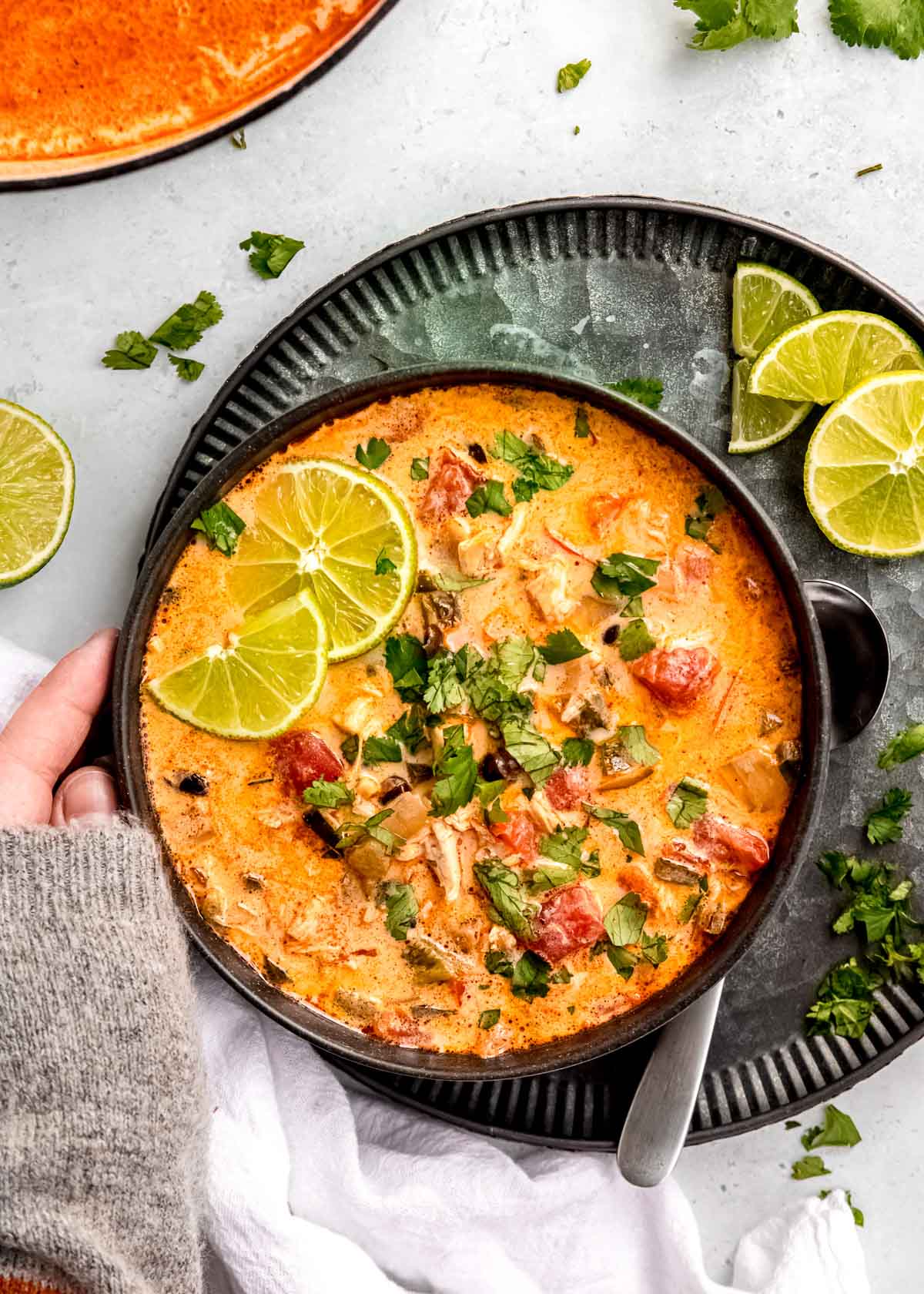 a hand touching a bowl of creamy poblano soup with tomatoes, beans, and chicken; spoon beside bowl on a dark charger with additional lime slices