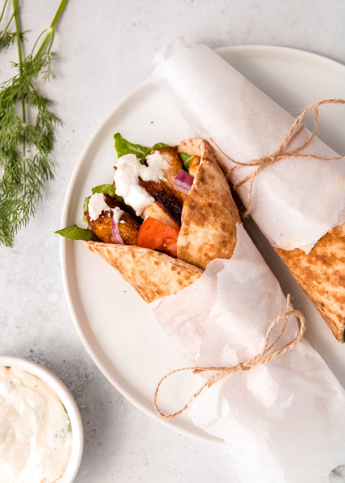 overhead shot showing two chicken shawarma pita wraps with veggies on a white plate with a side of creamy yogurt sauce