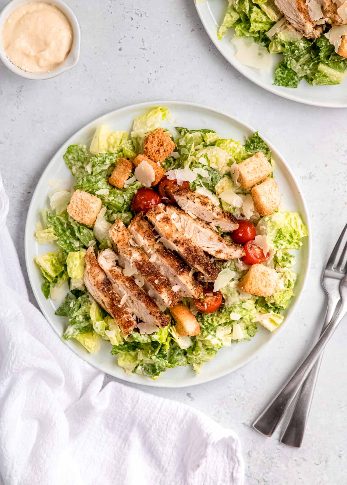 overhead image of grilled chicken caesar salad on white plate with fork and napkin on table
