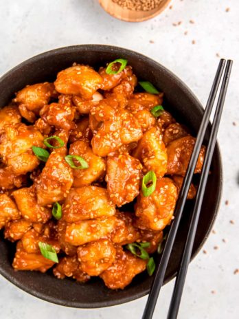 close up overhead image of sesame chicken and chopsticks in black bowl