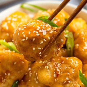 close up image of sweet and sour chicken being held between chopsticks over white bowl