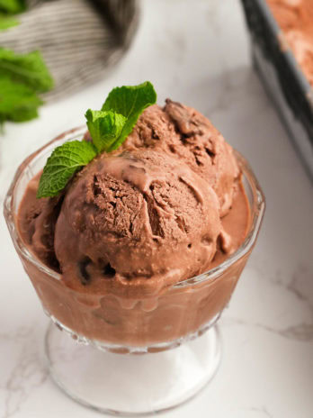 Three scoops of chocolate mint ice cream in a glass bowl garnished with peppermint leaves with a loaf pan full of ice cream in the background