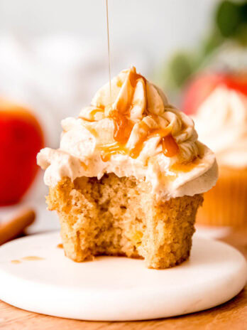 a gluten-free apple cinnamon cupcake on a white plate topped with a tall swirl of caramel buttercream being drizzled with extra caramel sauce; a bite has been bitten out of the cupcake to show the fluffy inside.