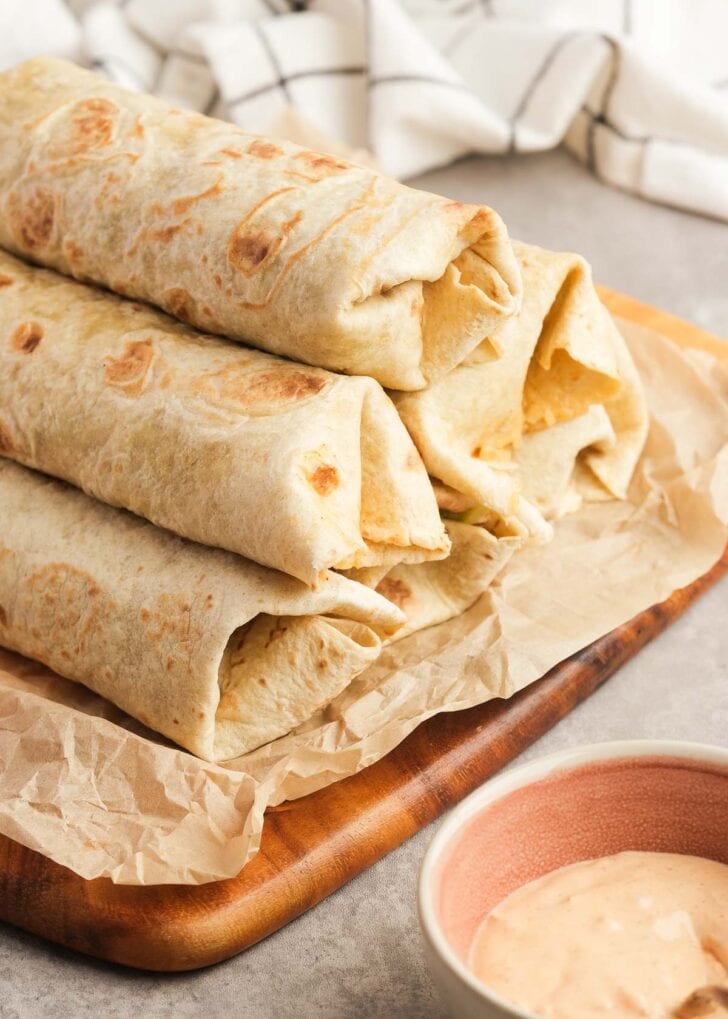 six folded and wrapped big mac wraps stacked on parchment paper beside a bowl of extra big mac sauce