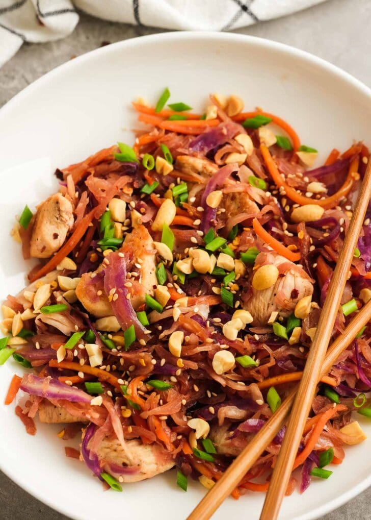 chopsticks crossed over chicken stir fry with purple cabbage, carrots, and a crunchy garnish of green onions and nuts