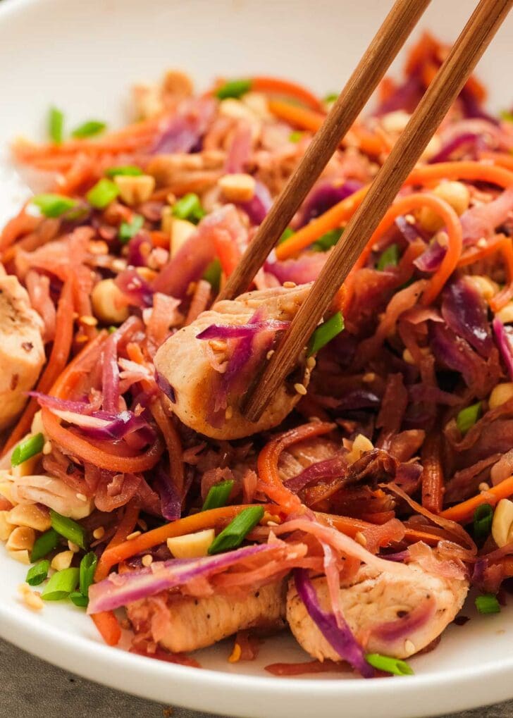 chopsticks holding a bite of chicken in a white dish full of egg roll in a bowl