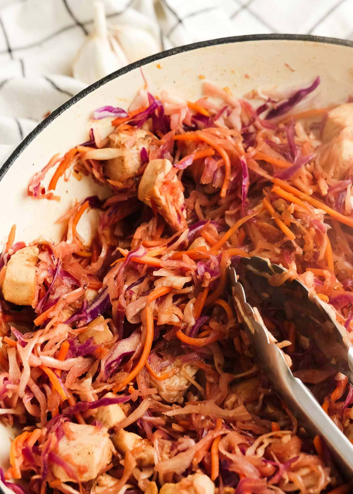 a pair of tongs tossing the chicken, cabbage, and carrots in a skillet with egg roll sauce