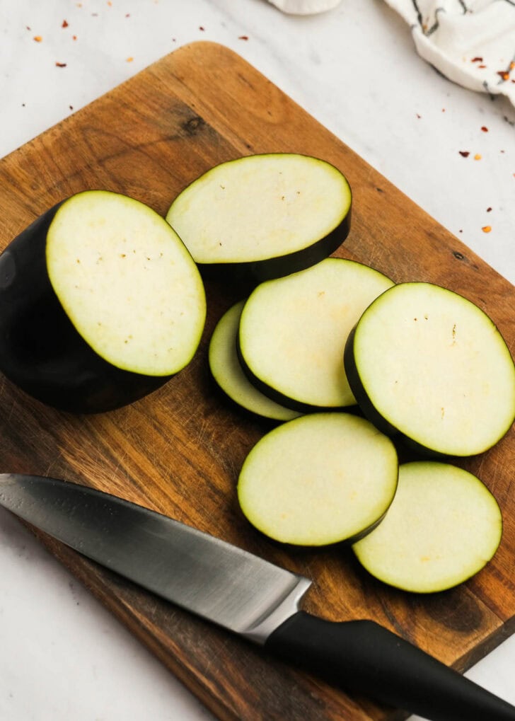 eggplants sliced into 1/2-inch thick rounds on a cutting board with a knife.