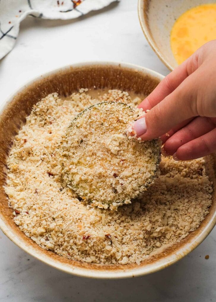 A slice of eggplant fully coated in seasoned breading after dipping into a bowl of eggs and a bowl of breadcrumbs, parmesan, and seasonings.