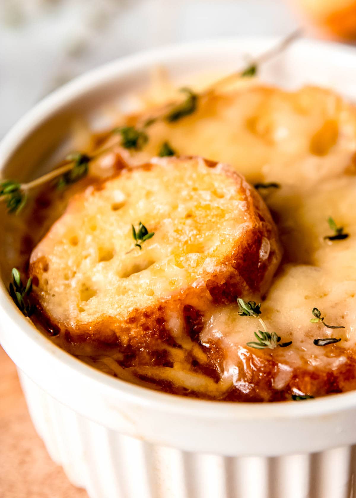 closeup view of a white ramekin full of French Onion Chicken Soup topped with melty gruyere, toasted baguette, and fresh thyme