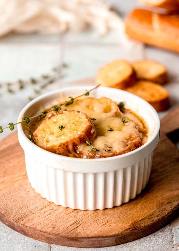 side shot of ramekin full of french onion chicken soup on a cutting board in front of extra baguettes and sprigs of thyme