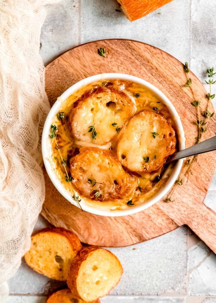 overhead shot showing a white ramekin full of french onion chicken soup topped with toasted bread and shredded gruyere cheese; spoon in ramekin, bowl on wooden cutting board, surrounded by thyme sprigs and more sliced baguette