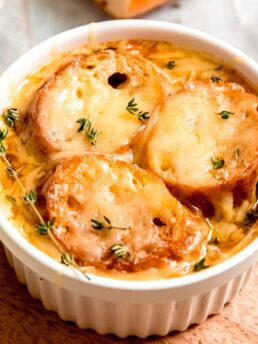 overhead closeup view of white ramekin filled to the brim with french onion chicken soup, toasted baguettes, and melted gruyere cheese