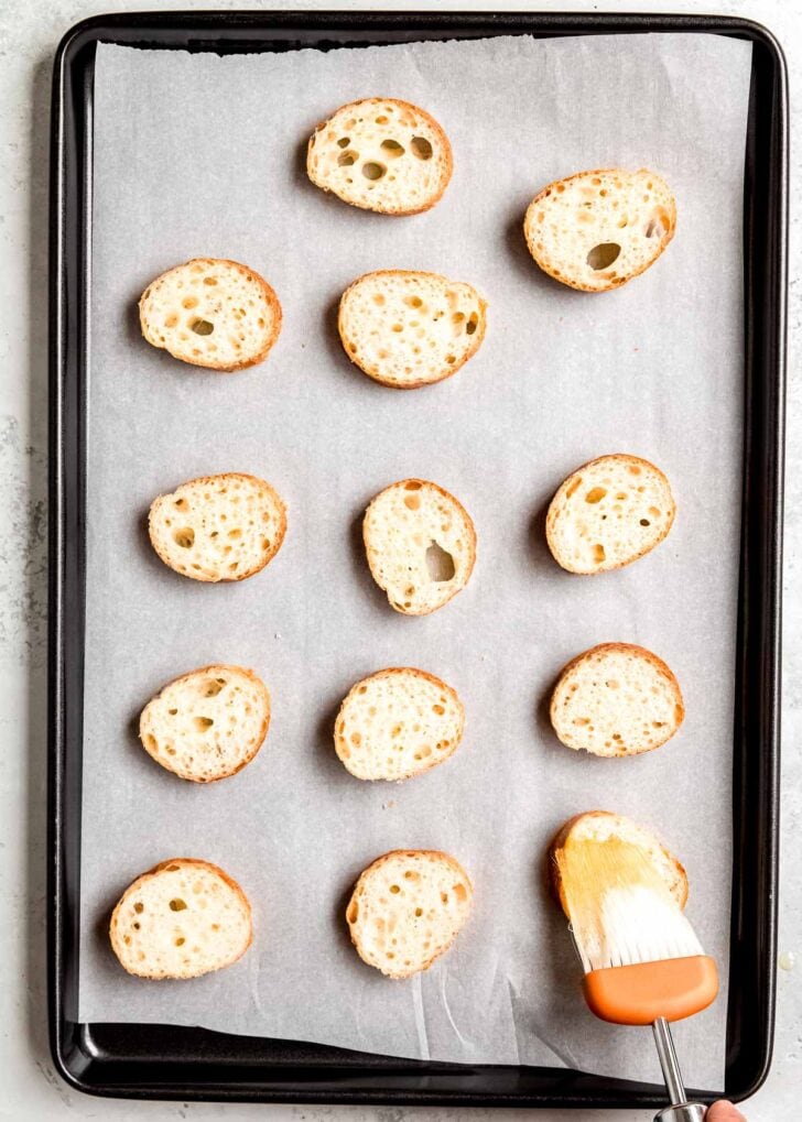 slices of french baguette on a parchment paper lined baking sheet. a culinary brush applying avocado oil to eat slice