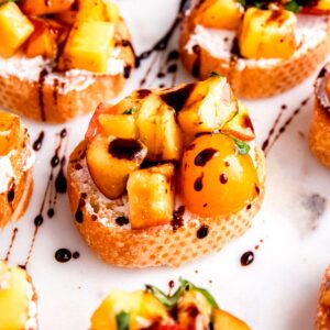 several pieces of peach bruschetta with goat cheese, peaches, tomatoes, basil, and balsamic reduction on a white platter