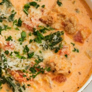 a white bowl full of creamy sausage and spinach soup topped with parmesan and chopped herbs