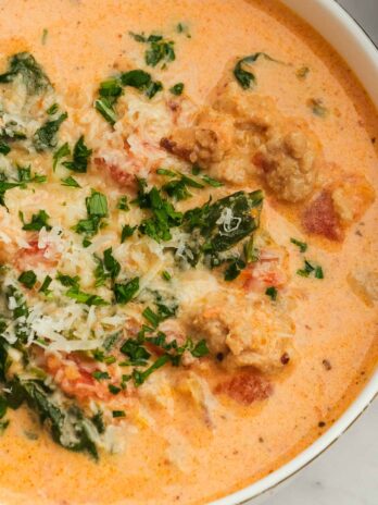 a white bowl full of creamy sausage and spinach soup topped with parmesan and chopped herbs