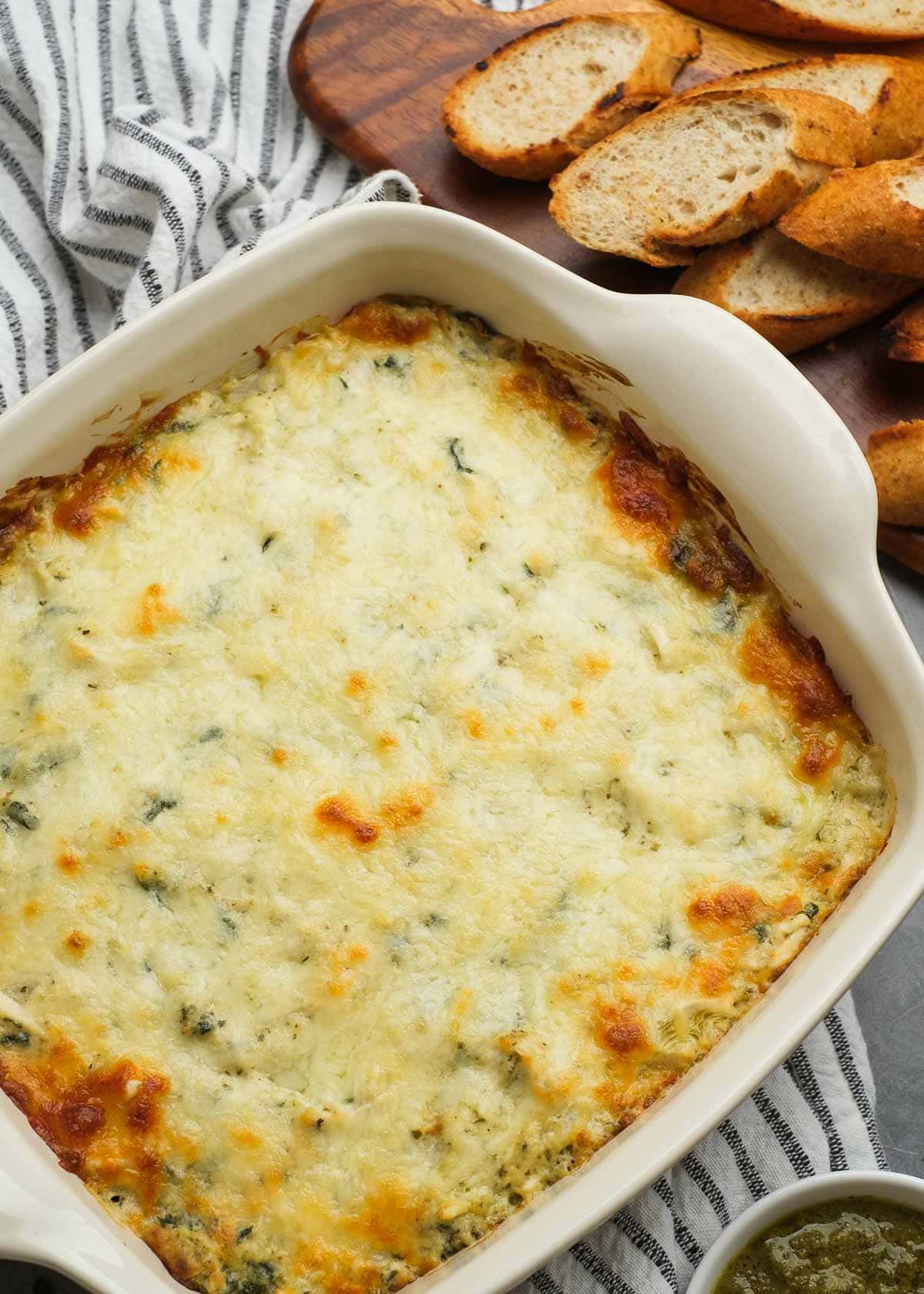 overhead shot showing a white baking dish filled with baked pesto chicken dip with a side of toasted baguette slices