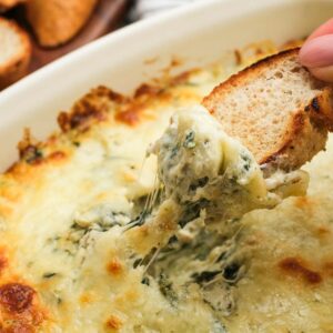 a hand dipping a piece of toasted bread into a creamy pesto chicken dip full of cheese, artichokes, and spinach