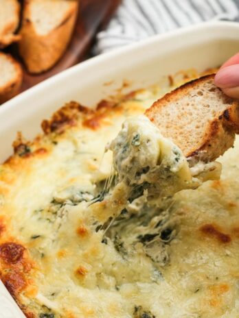 a hand dipping a piece of toasted bread into a creamy pesto chicken dip full of cheese, artichokes, and spinach