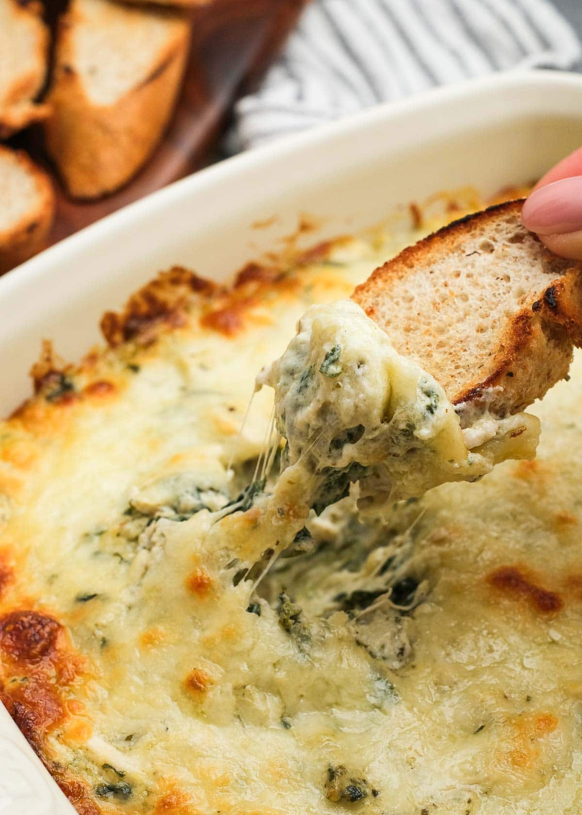 a hand dipping a piece of toasted bread into a creamy pesto chicken dip full of cheese, artichokes, and spinach