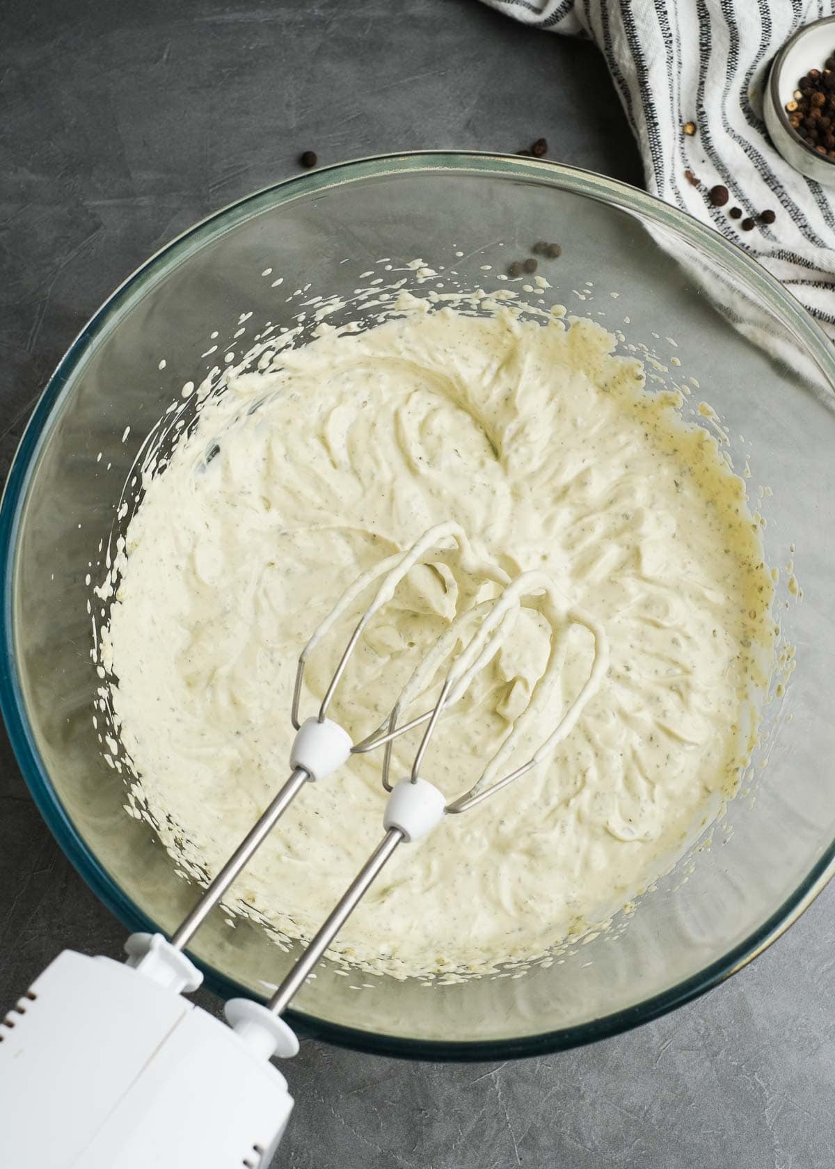 mayonnaise, greek yogurt, pesto, and cream cheese mixed together in a glass bowl with an electric hand mixer