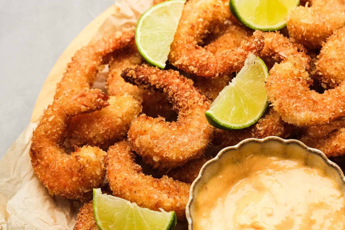 horizontal image showing crispy popcorn shrimp coated with panko and spices, tossed with lime wedges and served with a creamy sriracha sauce