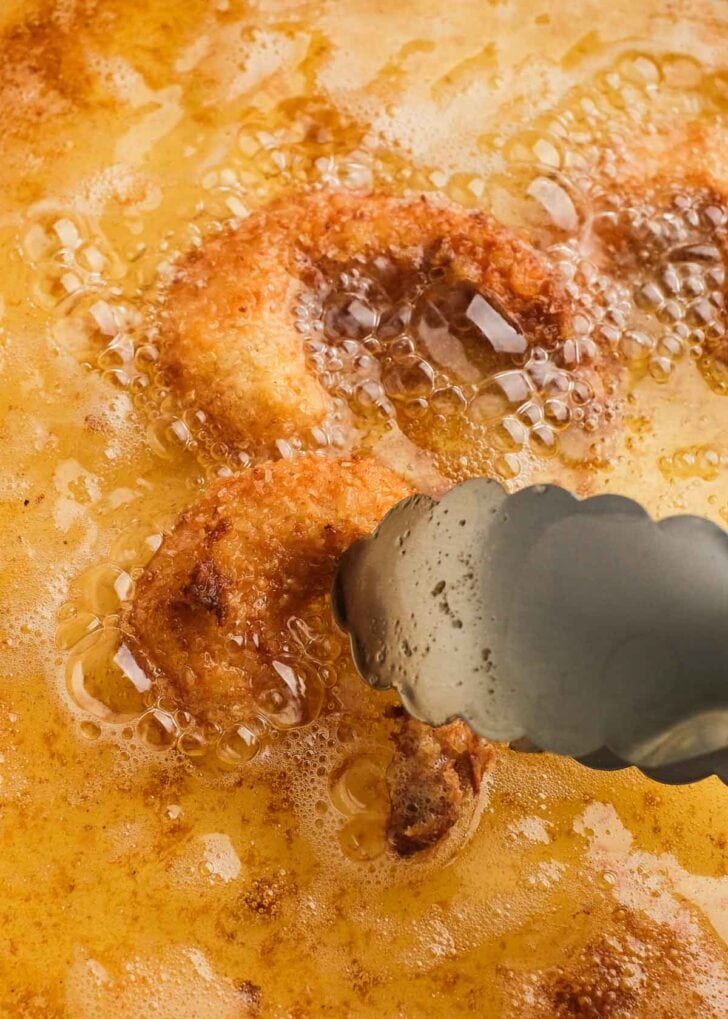 tongs flipping a piece of fried popcorn shrimp in oil