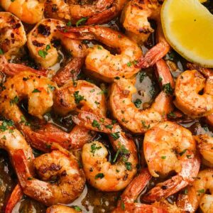 juicy shrimp covered in garlic butter sauce in a pan, garnished with fresh herbs and lemon wedges on the side