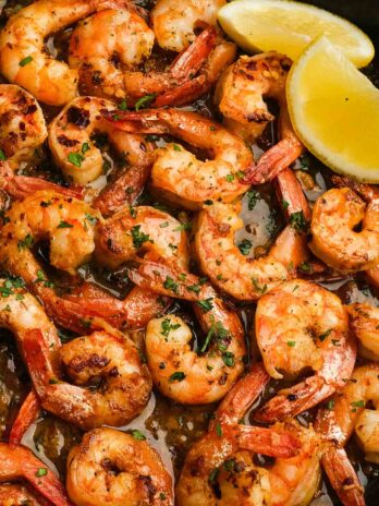 juicy shrimp covered in garlic butter sauce in a pan, garnished with fresh herbs and lemon wedges on the side