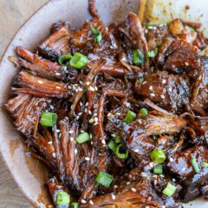 A bowl full of slow cooker shredded beef smothered in a sweet and spicy sauce and garnished with sesame seeds and green onions