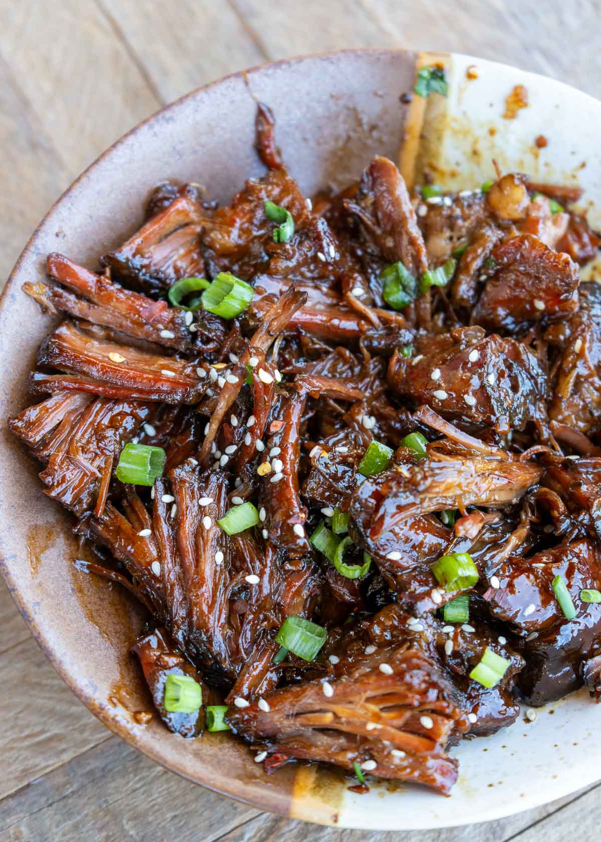 A bowl full of slow cooker shredded beef smothered in a sweet and spicy sauce and garnished with sesame seeds and green onions