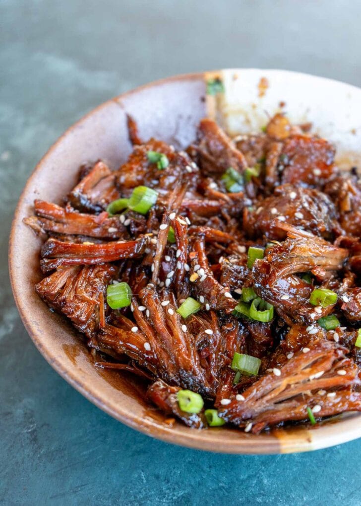 a bowl filled with juicy sweet and spicy shredded beef and garnished with chopped scallions and sesame seeds