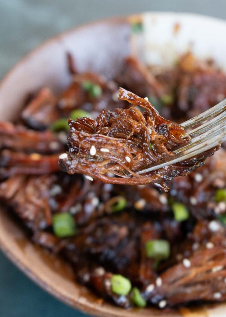 A fork holding up a big bite of juicy slow cooker shredded beef cooked in a sweet and spicy sauce