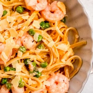 closeup view of a white bowl full of shrimp pasta with peas and a creamy sauce, garnished with shaved parmesan