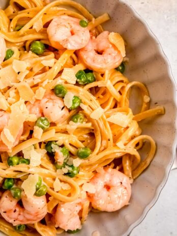 closeup view of a white bowl full of shrimp pasta with peas and a creamy sauce, garnished with shaved parmesan