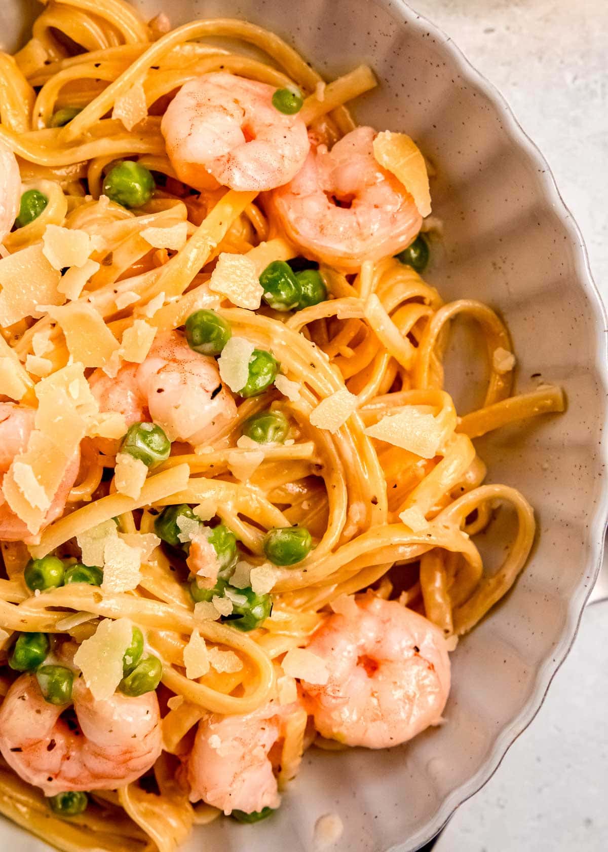 closeup view of a white bowl full of shrimp pasta with peas and a creamy sauce, garnished with shaved parmesan