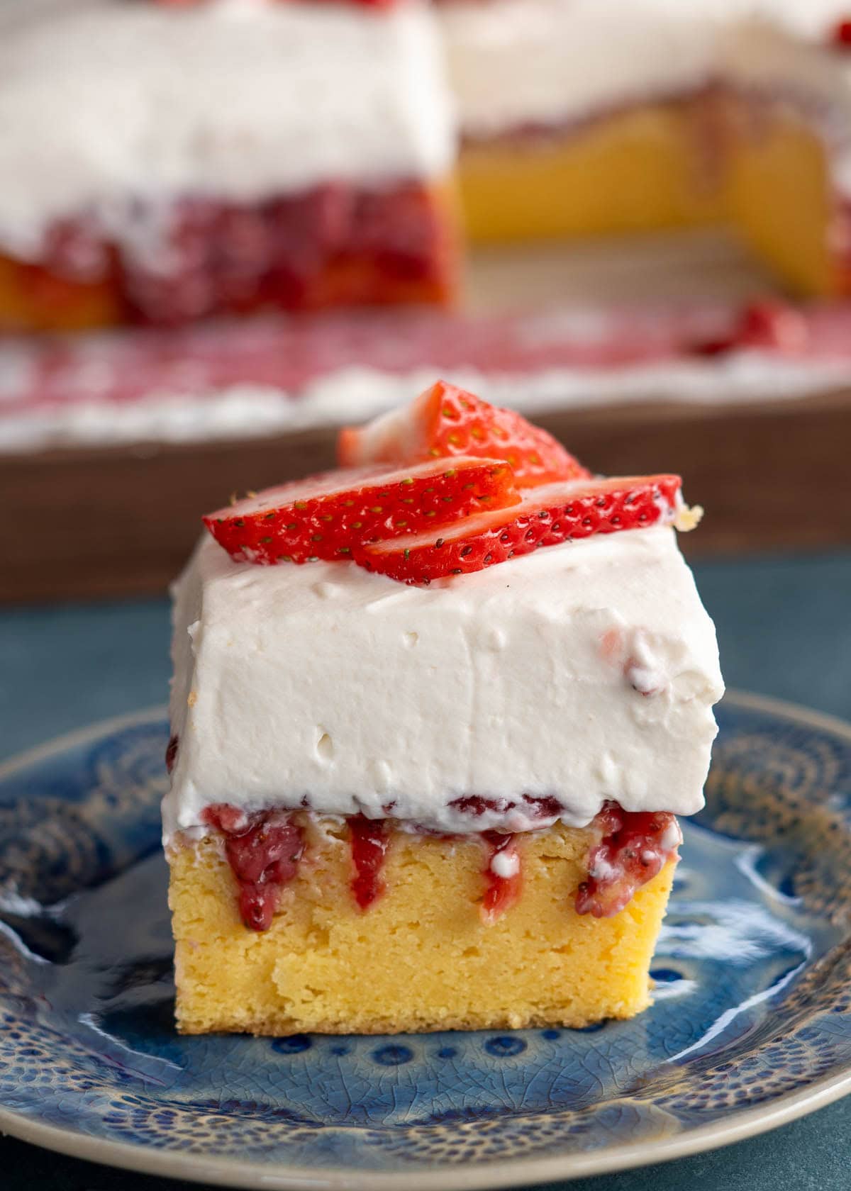 a piece of strawberry poke cake on a blue plate in front of the rest of the gluten-free cake